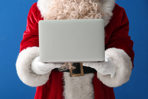 Santa Claus celebración de la computadora portátil moderna sobre fondo azul, primer plano - Foto, imagen