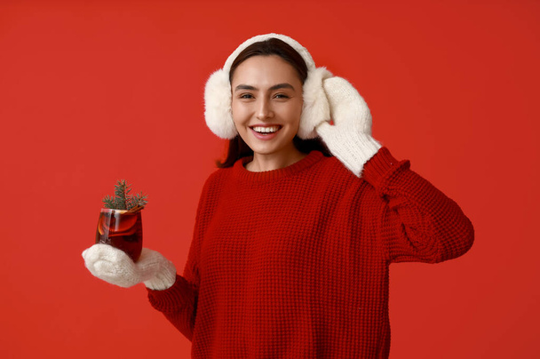 Happy young woman in warm mittens with glass of hot mulled wine on red background - Photo, Image