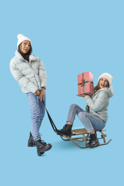 Mujeres jóvenes en ropa de invierno con regalo de Navidad trineo sobre fondo azul - Foto, imagen