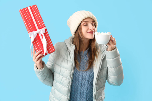 Young woman in winter clothes with cocoa and Christmas gift on blue background - Photo, Image