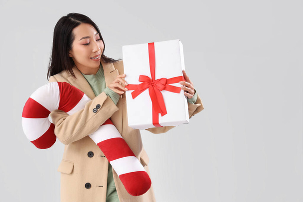 Young Asian woman with Christmas gift and candy cane pillow on light background - Photo, Image