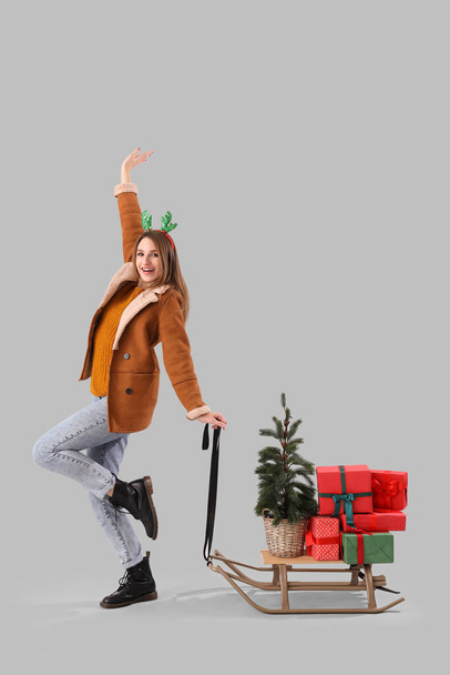 Junge Frau in Rentierhörnern mit Weihnachtsbaum und Geschenken auf Schlitten vor hellem Hintergrund - Foto, Bild