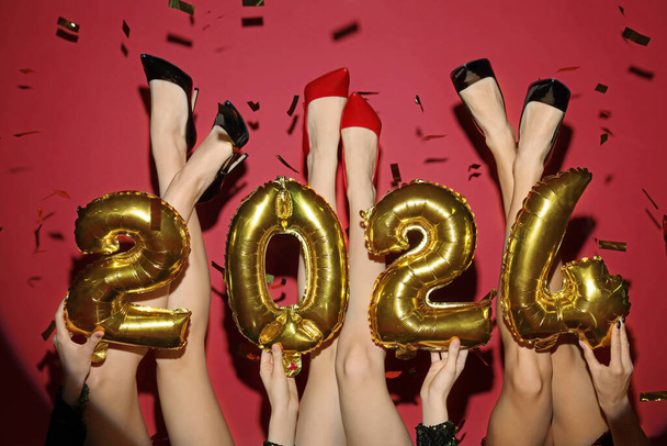 Ноги молодых женщин с рисунком 2024 и конфетти на красном фоне. Новогодний праздник - Фото, изображение