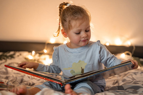 Niña leyendo pop-up libro en la cama de casa en el ambiente de Navidad con luces de fondo. Foto de alta calidad - Foto, imagen