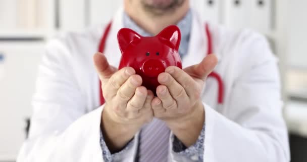 Young smiling male doctor holds piggy bank in hands. Medical insurance concept - Footage, Video