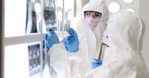 Dos doctores en trajes de protección y máscaras examinando imágenes de rayos X de pacientes. Neumonía viral de los pulmones concepto - Imágenes, Vídeo