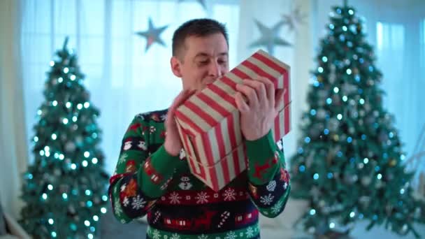 Retrato de um homem caucasiano a receber um presente de Natal. Happy sorrindo cara está feliz com a caixa perto da árvore de Natal. Imagens 4k de alta qualidade - Filmagem, Vídeo