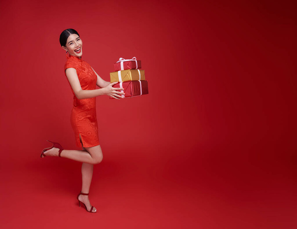 赤い伝統的な衣装のハッピー笑顔のアジアの女性は,赤い背景で隔離された中国の新年の祝福のためのギフトボックスを与えます. - 写真・画像