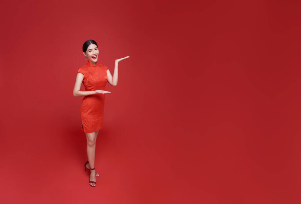 ハッピー中国の新年. 赤いコピースペースの背景に隔離された歓迎のジェスチャーで伝統的なチョンサム qipao ドレスを身に着けているアジアの女性. - 写真・画像