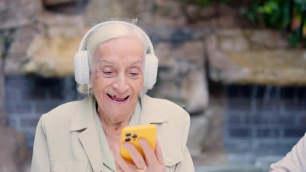 Vidéo d'une heureuse femme âgée écoutant de la musique avec des écouteurs et mobile à l'extérieur d'un gériatrique - Séquence, vidéo