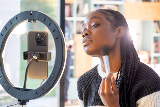 This image depicts a social media influencer woman engaged in her beauty routine, applying makeup with a brush in front of a ring light mirror that illuminates her face. Her expression is one of - Photo, Image