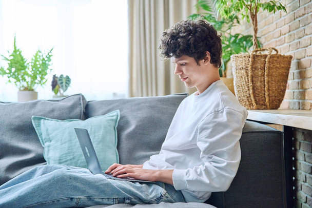 Jongeman tussen de 18 en 20 jaar, thuis, zittend op de bank in de woonkamer, laptop aan het typen. Internet online technologie, studie vrije tijd communicatie concept - Foto, afbeelding