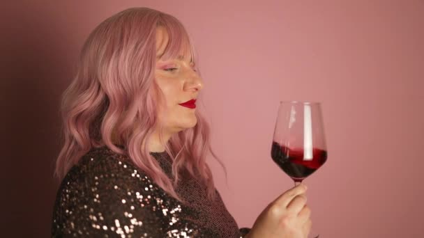 Mulher de cabelo rosa elegante positivo em vestido preto moderno brilhante segurando copo de vinho e degustação de bebida em pano de fundo rosa. Senhora comemorando um aniversário em uma festa. - Filmagem, Vídeo