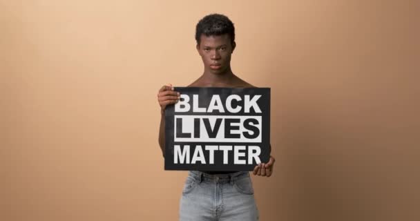 Молодой черный мужчина без рубашки с табличкой с фразой "ЧЕРНЫЙ ЖИВИТ МАТЕР" сердито смотрит на камеру в студии на бежевом фоне - Кадры, видео