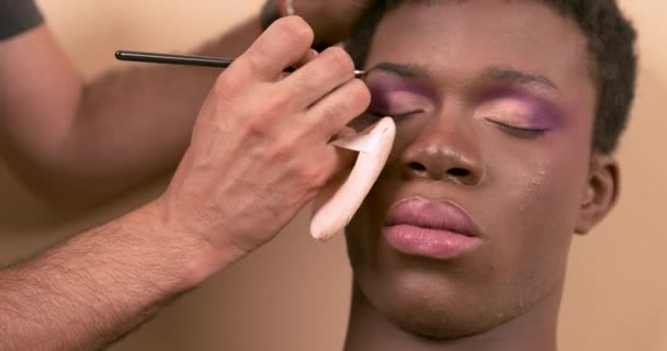 Crop visagiste masculino anónimo haciendo maquillaje de cejas en drag queen afroamericano mientras se prepara para la sesión de fotos en el estudio de luz - Imágenes, Vídeo