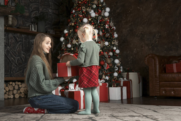 La madre joven y su hija pequeña abren el regalo de Navidad, la decoración de año nuevo en el interior. Mamá e hija cerca del árbol de Navidad. Nochebuena. - Foto, imagen