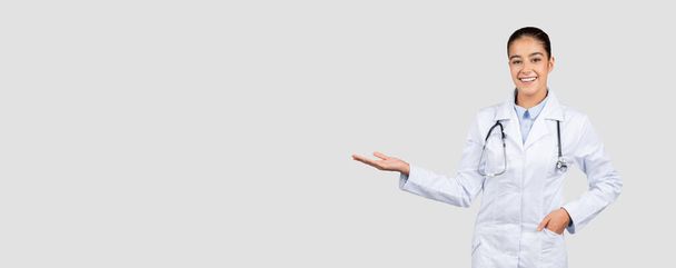 Χαρούμενη Ευρωπαίος γιατρός χιλιετηρίδας σε λευκό παλτό, με στηθοσκόπιο κρατήσει αντίγραφο χώρο στο χέρι, απομονώνονται σε γκρι φόντο στούντιο, πανόραμα. Ιατρική διαφήμιση και προσφορά, παρουσίαση υγειονομικής περίθαλψης, πώληση - Φωτογραφία, εικόνα