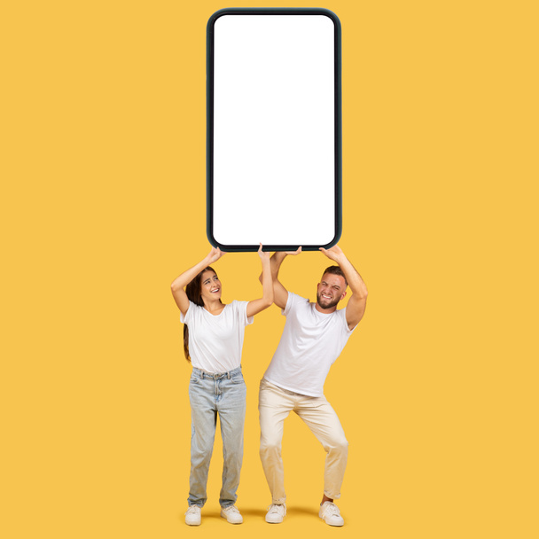 Feliz divertida familia europea milenaria en camisetas blancas sostienen enorme teléfono inteligente con pantalla vacía, aislado en el fondo del estudio amarillo. Compras en línea, aplicación, sitio web de venta, anuncio y oferta - Foto, imagen