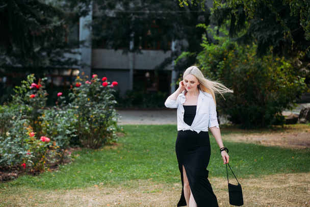Une belle femme mince et souriante aux longs cheveux blonds, en robe longue noire et chemise blanche, se promène dans le parc sur fond de buissons fleuris de la ville. - Photo, image