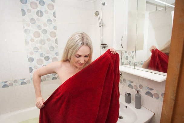 Блондинка заворачивается в красное полотенце, в ванной. Творческий образ на тему здоровья, гигиены, заботы о своем теле. - Фото, изображение