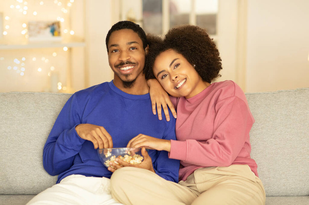 Χαλαρωμένο μαύρο ζευγάρι παρακολουθεί ταινία στην τηλεόραση απολαμβάνοντας ρομαντική βραδιά στο σπίτι, τρώγοντας ποπ κορν και αγκαλιάζοντας τον καναπέ, χαλαρώνοντας στην άνεση του οικιακού τους περιβάλλοντος το Σαββατοκύριακο - Φωτογραφία, εικόνα