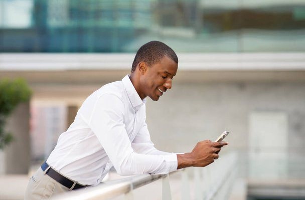 Lächelnder schwarzer Geschäftsmann, der sich an Geländer lehnt und sein Smartphone im Freien benutzt, junger afroamerikanischer männlicher Unternehmer, der sein Handy im Freien benutzt, Online-Nachrichten auf dem Handy, Kopierraum - Foto, Bild
