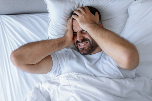 Een slapeloze man met een baard die alleen in bed ligt en thuis pijn in zijn hoofd doet, lijdt aan migraine en slapeloosheid en zware gedachten heeft. Problemen met slapen. Bovenaanzicht opname - Foto, afbeelding