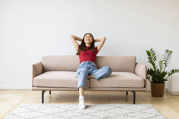 Home Comfort. Porträt einer lächelnden jungen Asiatin, die sich auf der Couch zurücklehnt, einer glücklichen Millennial Lady, die sich auf dem Sofa im Wohnzimmer ausruht, mit geschlossenen Augen und Händen hinter dem Rücken entspannt, Kopierraum - Foto, Bild