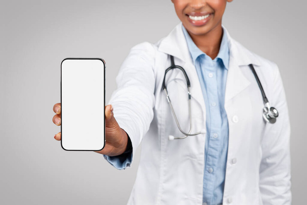 Рада, что чёрная юная леди доктор в белом халате показала телефон с пустым экраном, изолированный на сером фоне студии. Медицинские услуги, сайт объявлений и предложений, рекомендации приложений - Фото, изображение