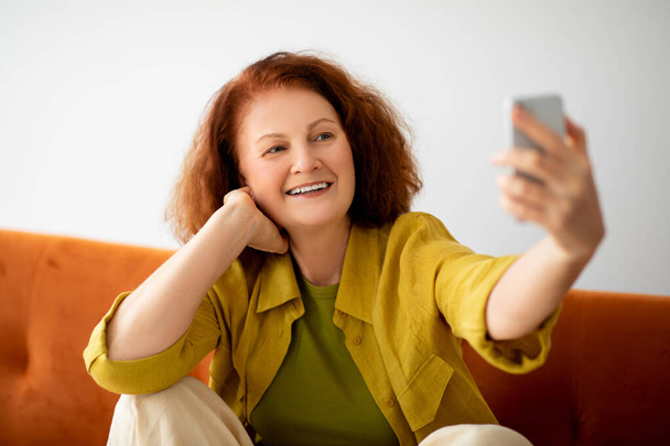 Szczęśliwa ruda starsza kobieta robi sobie selfie na smartfonie siedząc na kanapie w domu, radosna, doświadczona technicznie starsza pani korzystająca z telefonu komórkowego, robiąca zdjęcia i dobrze się bawiąca, ujęcie zbliżeniowe - Zdjęcie, obraz