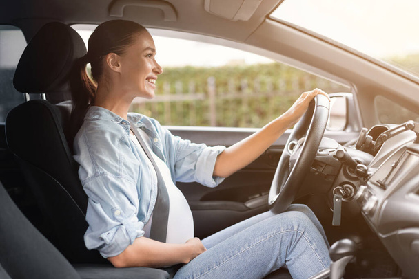 Ciężarna młoda dama siedzi w samochodzie delikatnie dotykając brzucha, uśmiechając się jadąc przez miasto i myśląc o przyszłym dziecku i macierzyństwie. Ciąża i bezpieczna jazda samochodem, ubezpieczenie - Zdjęcie, obraz