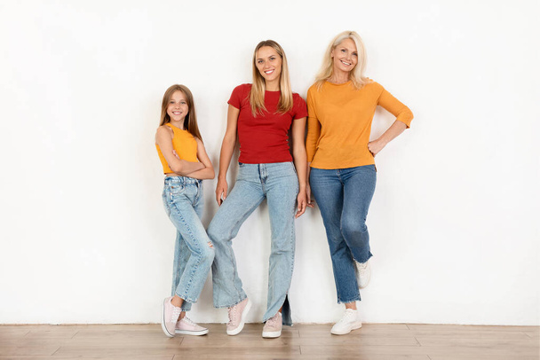 Onnellinen kaunis kolmen sukupolven naisten perhe poseeraa yhdessä valkoisen seinän takana, hymyillen iloisesti kameralle, täyspitkä. Äiti, tytär ja mummo rennoissa vaatteissa - Valokuva, kuva