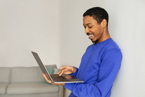 Allegro uomo afroamericano che tiene il computer portatile e lavora online tramite computer appoggiato sulla parete bianca, in piedi a casa, vista laterale. Giovani Freelancer Websurfing, Pubblicità Sito Web - Foto, immagini