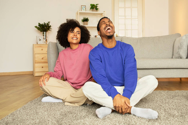 Весёлые чёрные муж и жена сидят на полу и смеются в современной гостиной дома. Молодые супруги наслаждаются выходными вместе в домашней обстановке, выражая положительные эмоции - Фото, изображение