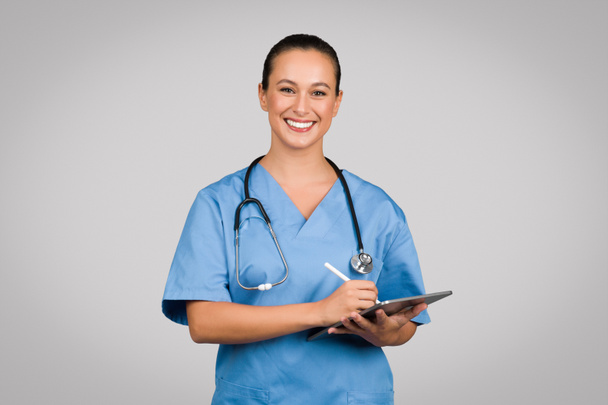 Φιλική γυναίκα νοσοκόμα σε μπλε τρίβει κρατώντας ψηφιακό δισκίο, ενσωματώνοντας σύγχρονη τεχνολογία υγειονομικής περίθαλψης με φωτεινό, προσιτό συμπεριφορά - Φωτογραφία, εικόνα