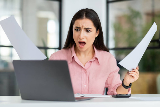 Verdrietig geschokt jonge Europese vrouw manager met open mond opstaan papieren, kijk naar laptop lijden aan werkproblemen, fout in het coworking office. Stress bij het bedrijfsleven, uiterste datum en druk - Foto, afbeelding