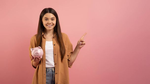 Usmívající se dospívající dívka drží prasátko banky v ruce a ukazuje stranou na kopírovací prostor, šťastná dospívající žena doporučuje nabídku, demonstrující volné místo pro reklamu, stojící na růžovém pozadí studia - Fotografie, Obrázek