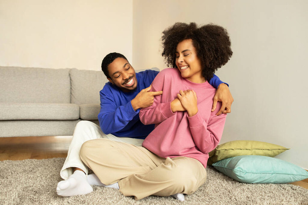 Boldog afro-amerikai férj és feleség közös pillanatai nevetés és ölelés, csiklandozzák egymást a kényelmes nappaliban otthon, szimbolizálja öröm és játékosság a romantikus kapcsolat - Fotó, kép