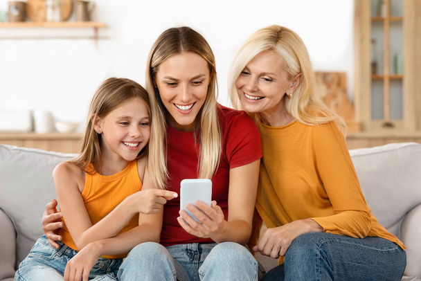 Tres generaciones en familia disfrutando del tiempo con un smartphone. Abuela, madre e hija sonriendo mientras miran un teléfono celular, compartiendo un momento de unión - Foto, imagen