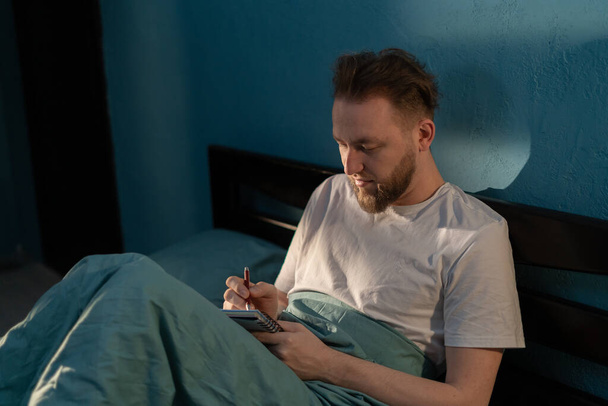 Hombre caucásico sentado en la cama, escribiendo en el diario, tomando notas en el diario por la noche. Copiar espacio - Foto, Imagen