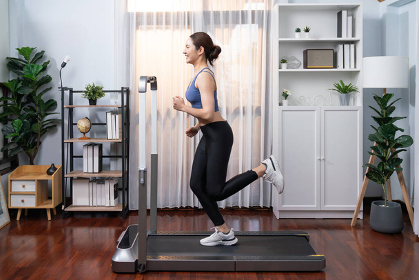 Full length side view of energetic and strong athletic asian woman running running machine at home. Pursuit of fit physique and commitment to healthy lifestyle with home workout and training. Vigorous - Photo, Image