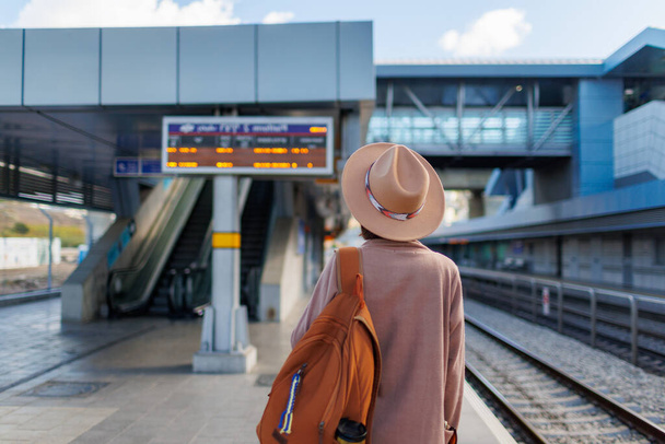 podróż pociągiem. Młoda kobieta w płaszczu i kapeluszu stoi na stacji i patrzy na rozkład jazdy pociągów. przygoda i podróże. - Zdjęcie, obraz