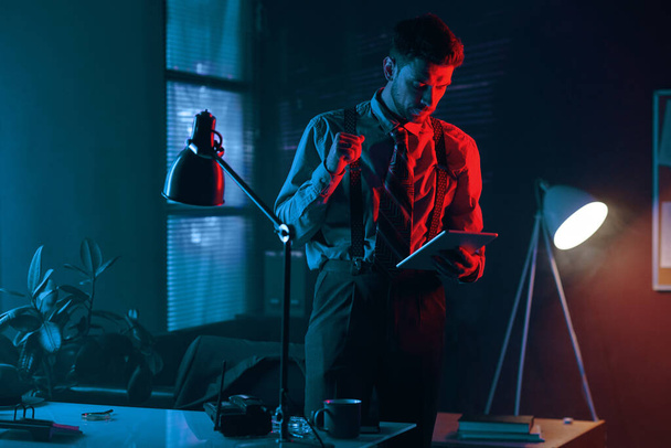 Junger eleganter Geschäftsmann mit Tablet blättert in elektronischem Finanzdokument und steht im dunklen Büro mit Neonlicht - Foto, Bild