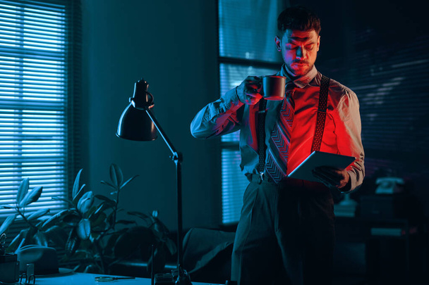 Nuori vakava liikemies, jolla on kuppi teetä tai kahvia tabletin avulla, kun hän seisoo pimeässä toimistossa myöhään yöllä ja lukee sähköistä asiakirjaa - Valokuva, kuva