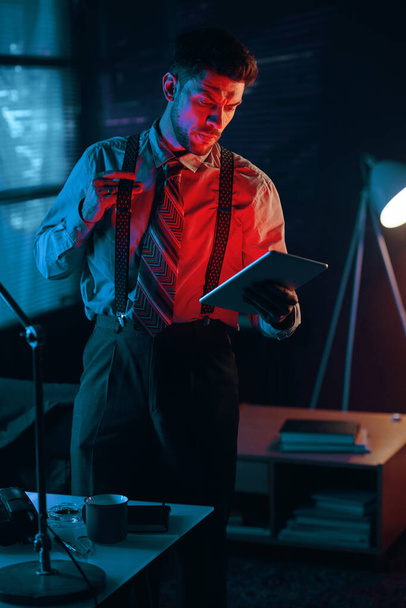 Young serious male analyst or director of business company with tablet looking through data in electronic document in dark office - Photo, Image