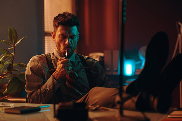 Jonge zakenman met zijn benen op tafel die een sigaret opsteekt terwijl hij in donker kantoor zit en na overwerk pauze heeft - Foto, afbeelding