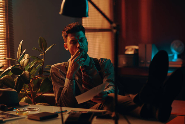 Junge männliche Büroangestellte rauchen Zigarette und betrachten Fotos, während sie am Arbeitsplatz im dunklen Büro sitzen und sich nach der Arbeit ausruhen - Foto, Bild