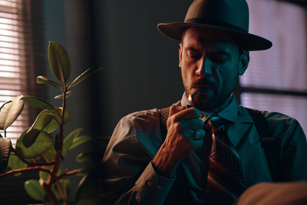 Молодой ретро-человек в шляпе и формальной одежде закуривает сигарету, сидя перед камерой с зеленым домашним растением справа. - Фото, изображение