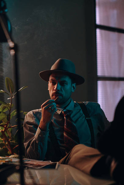 Junger nachdenklicher Retro-Mann mit Hut und Formalbekleidung hält Zigarette in der Hand und denkt an etwas, während er im dunklen Büro oder Zimmer sitzt - Foto, Bild