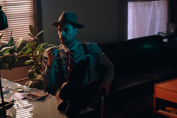 Νεαρός σοβαρός επιχειρηματίας με καπέλο και επίσημο ένδυμα κοιτάζοντας στην άκρη και καπνίζοντας τσιγάρο ενώ κάθεται στο χώρο εργασίας στο σκοτεινό γραφείο - Φωτογραφία, εικόνα